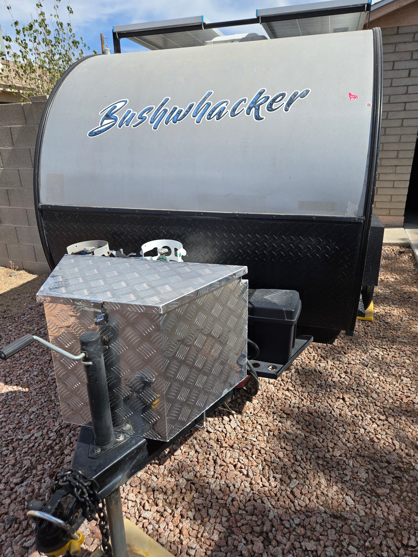 Front view of a Bushwhacker off-road teardrop trailer for rv rental, showing solar panels, front storage box, and gravel ground.