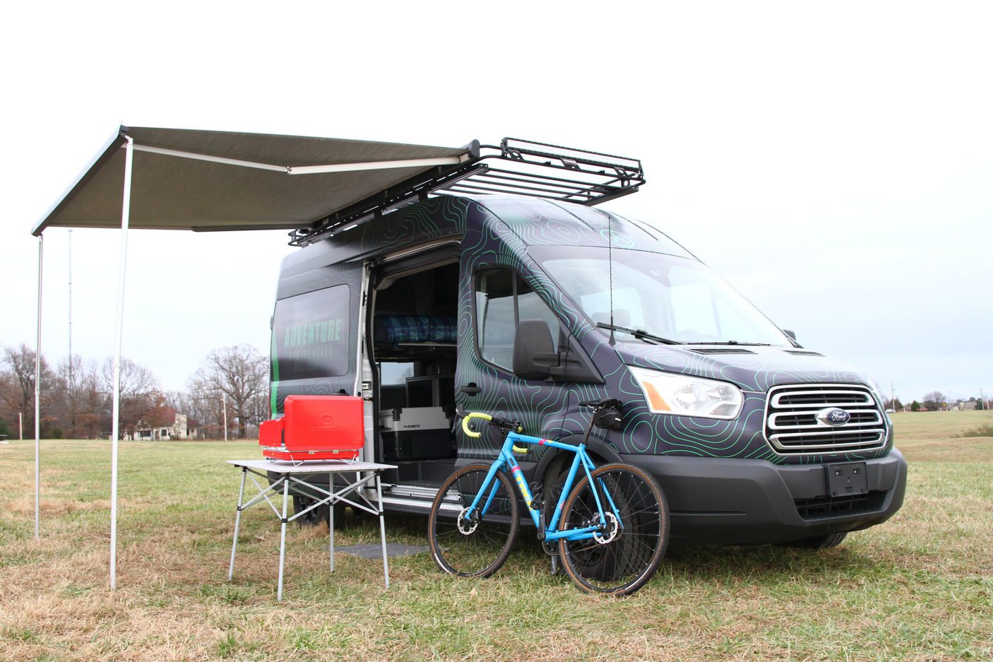 Adventure rv rental van, with graphic design, awning, bike, & camp setup in a field. Perfect for outdoor travel.
