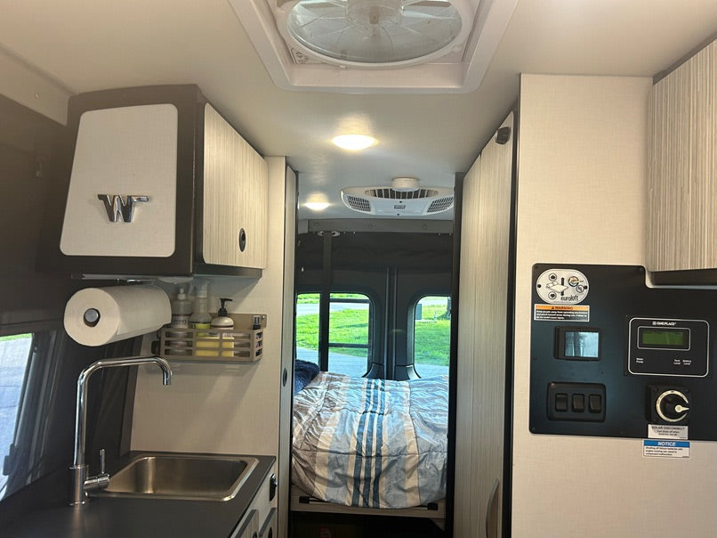 Inside an rv rental: kitchen with sink, overhead cabinets, and bed. Control panel on right. A compact mobile home.