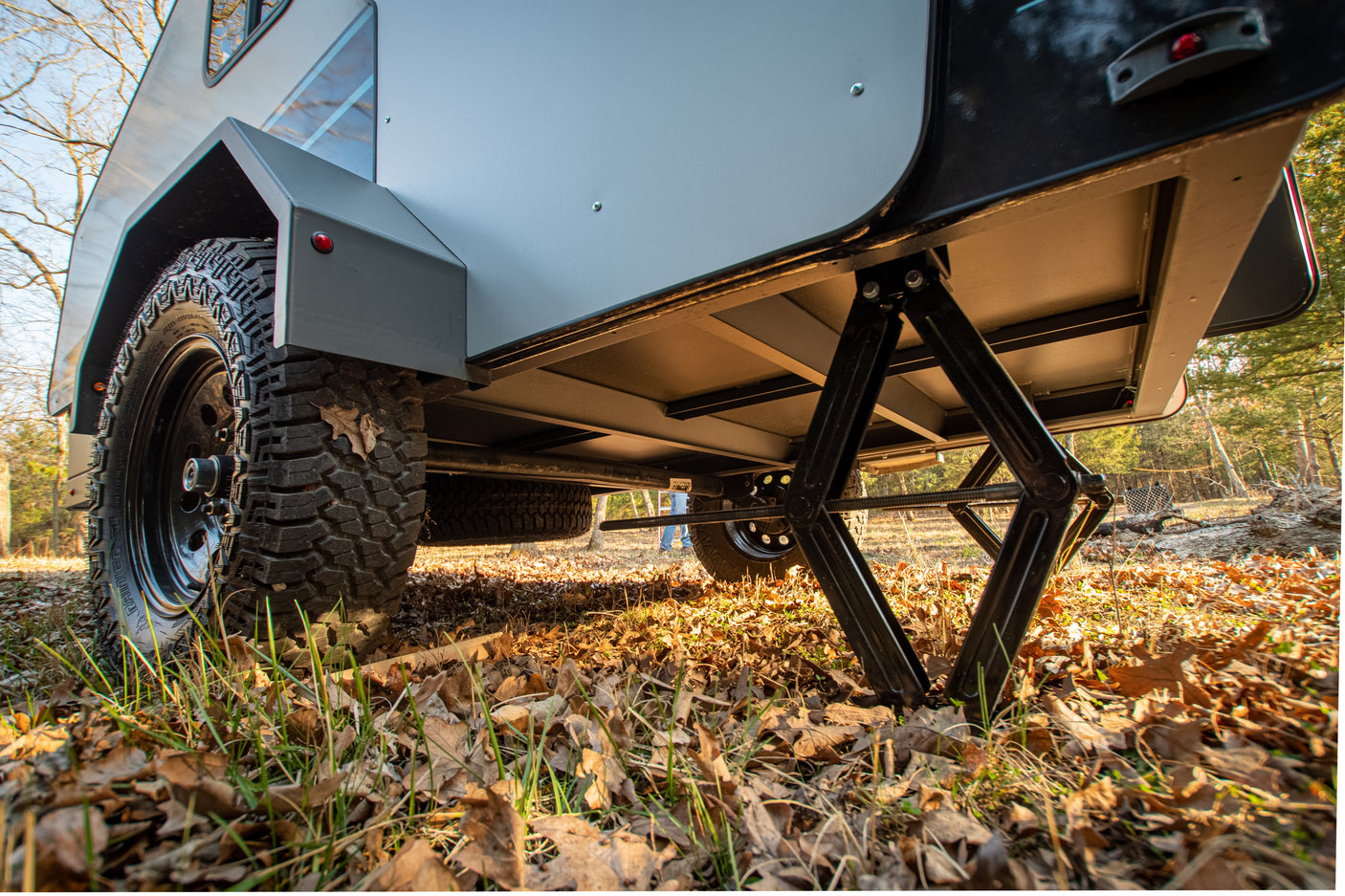 Low-angle view of an off-road travel trailer for RV rental, showcasing its rugged tire and stabilizing jack on autumn leaves.