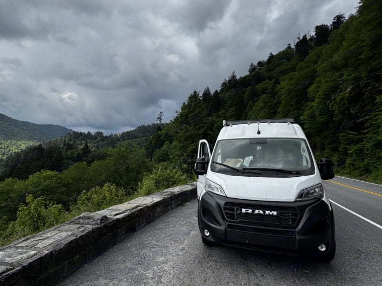 Our white Ram ProMaster RV rental on a mountain road, with cloudy skies and lush forests. Adventure awaits!
