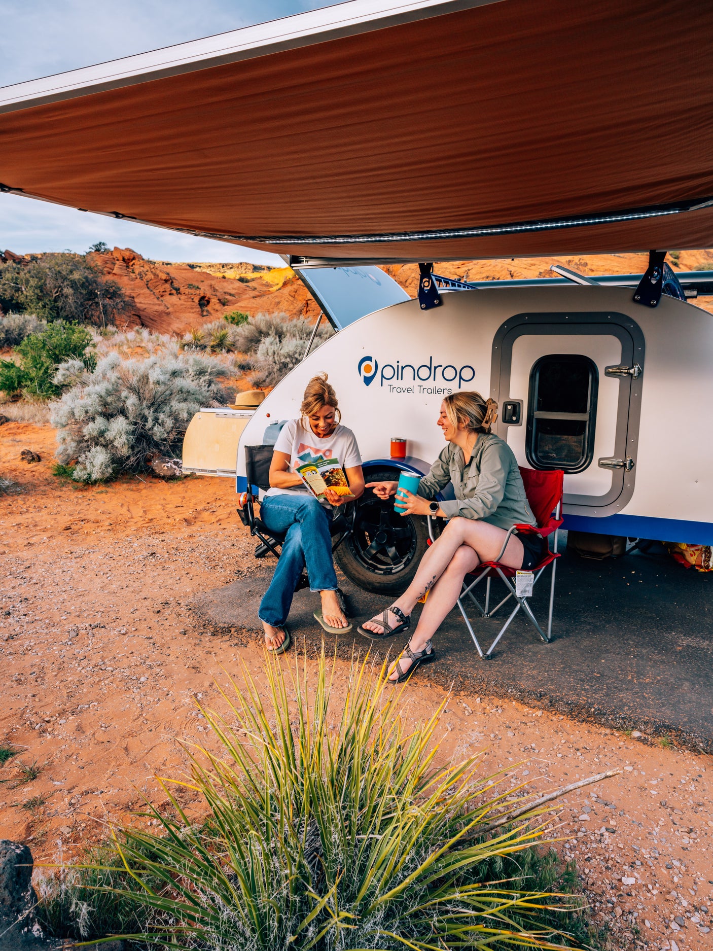 Two women relax under an awning next to a Pindrop rv rental travel trailer in the desert. One reads, the other sips a drink.