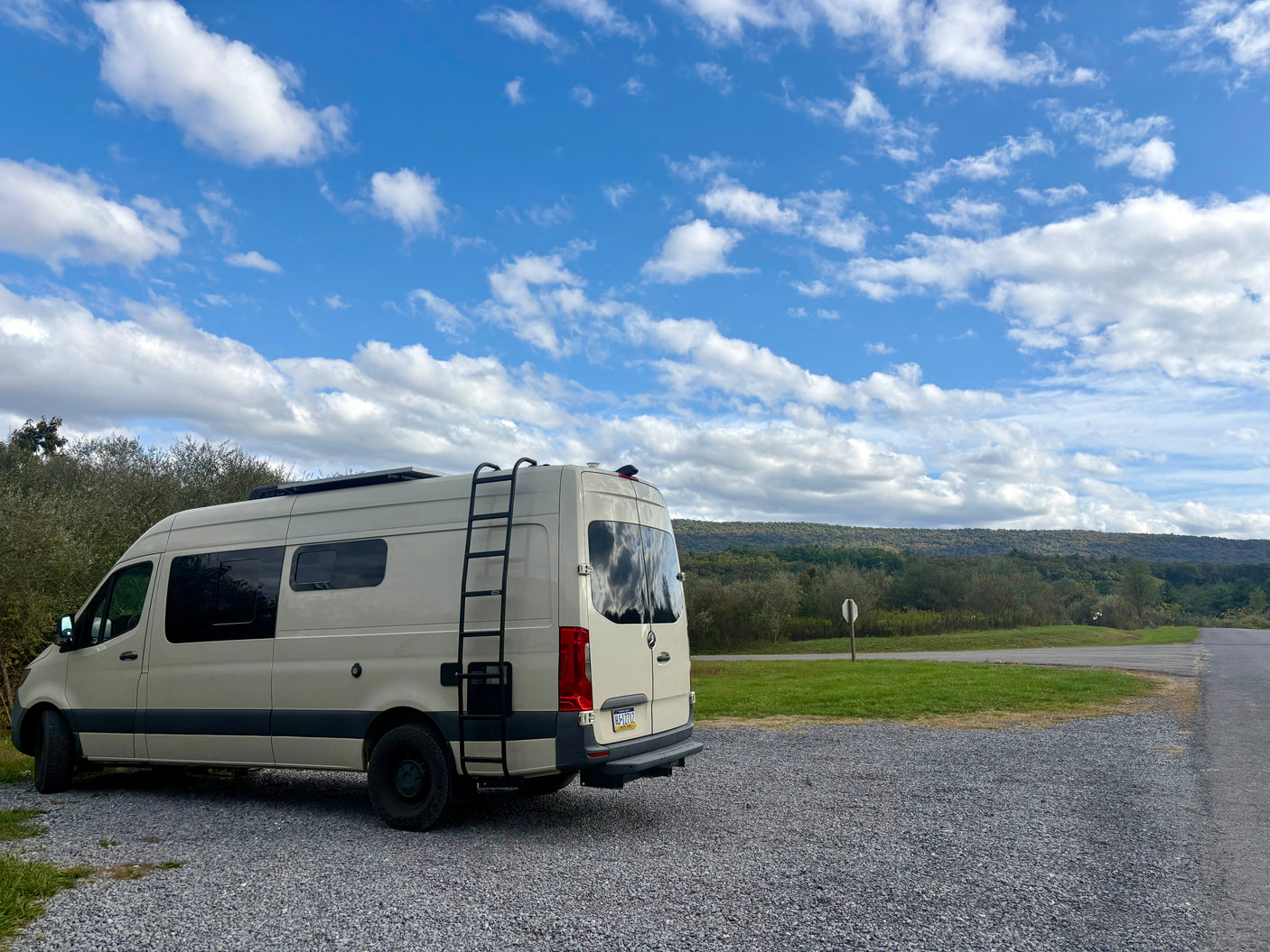 Light-colored Sprinter RV rental parked on gravel. Scenic view of green hills, trees, and blue sky with white clouds.