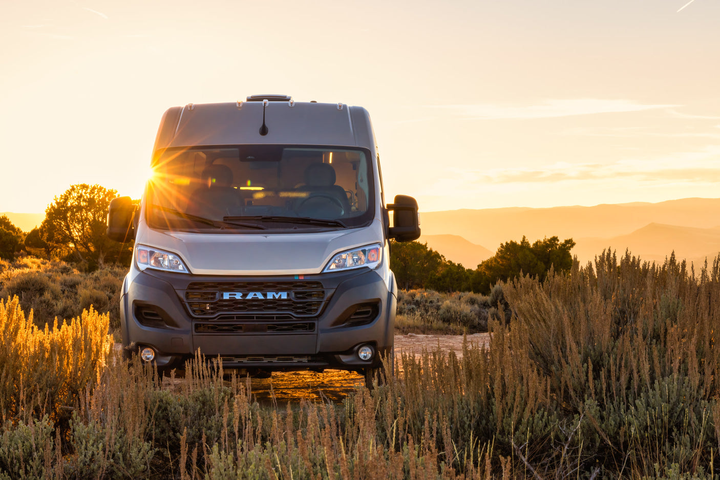 Light grey Ram ProMaster RV rental parked in golden desert brush at sunset, with sun rays peeking over the horizon.