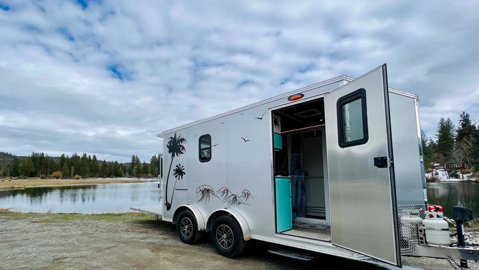 An open silver rv rental travel trailer with palm tree decals overlooks a calm lake and forest under a cloudy sky.