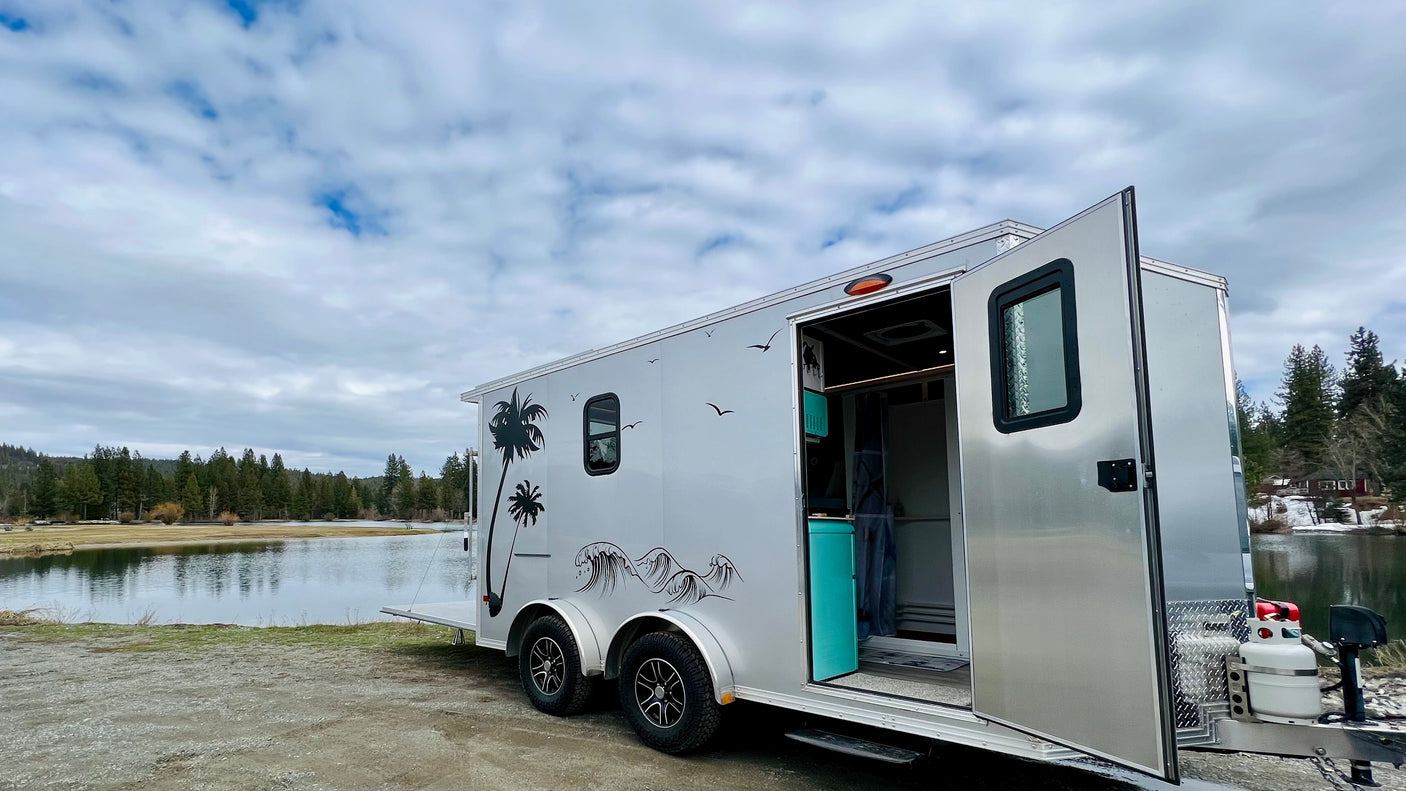 An open silver rv rental travel trailer with palm tree decals overlooks a calm lake and forest under a cloudy sky.