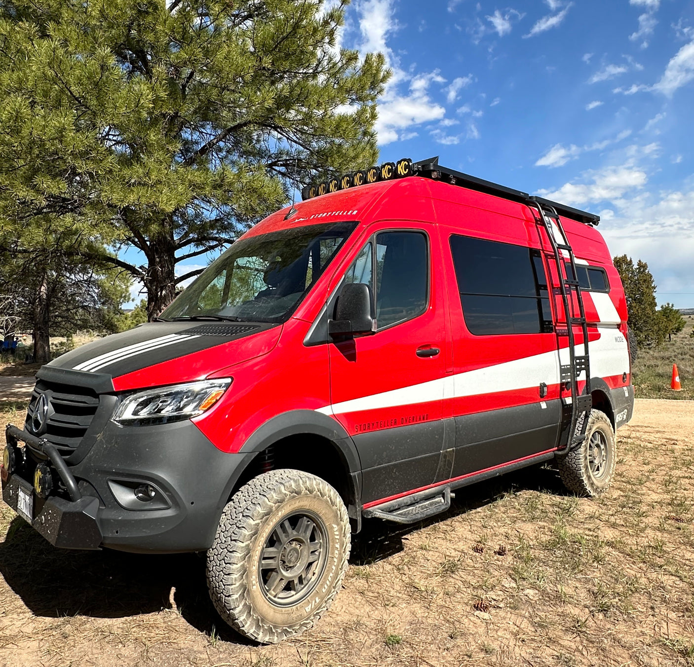 A vibrant red Storyteller Overland camper van, equipped for off-road adventures, parked among pine trees. Ideal for an RV rental.