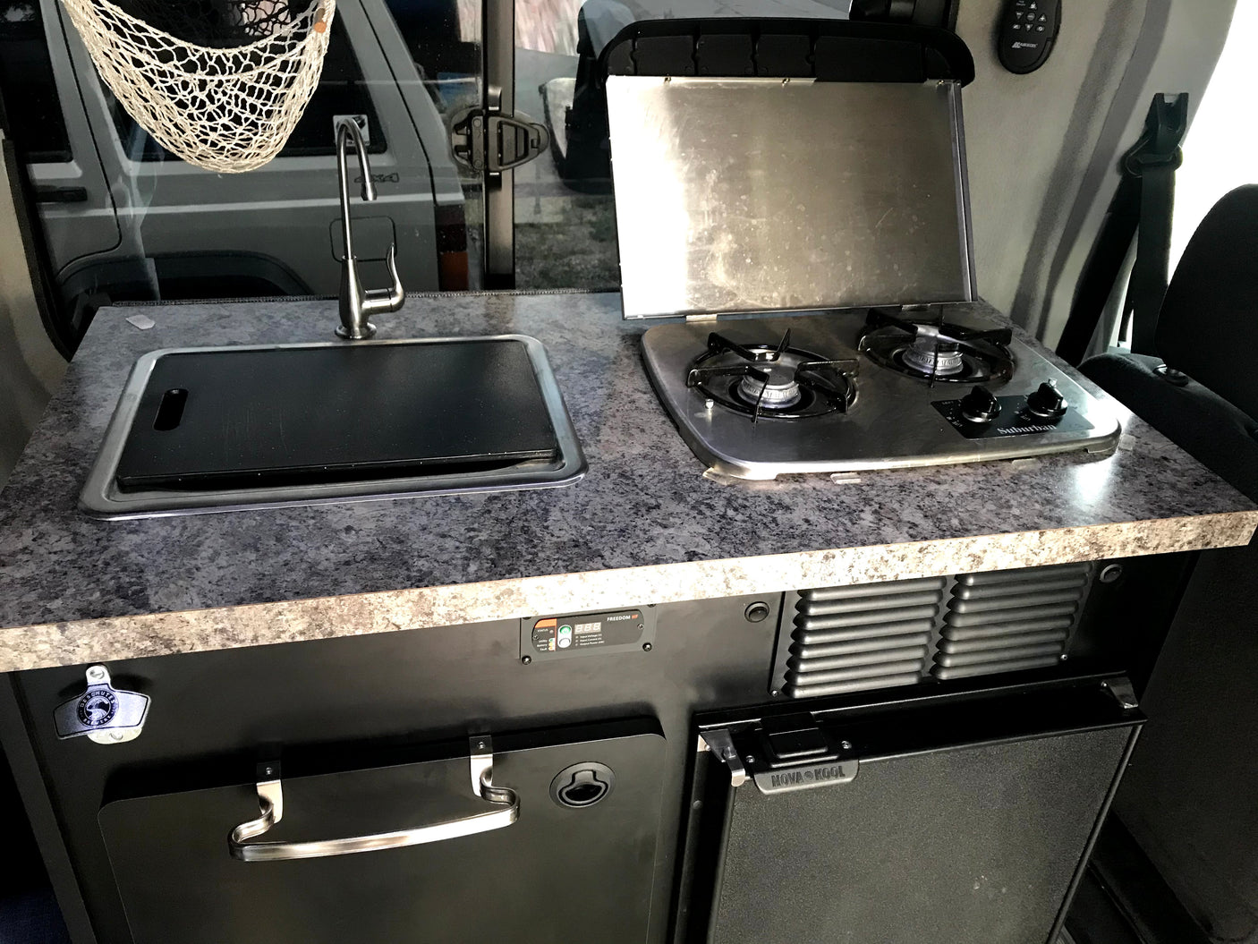 RV rental kitchen: a countertop with a sink (covered) and two-burner stove. Below, a Nova Kool fridge, pull-out table, and bottle opener.