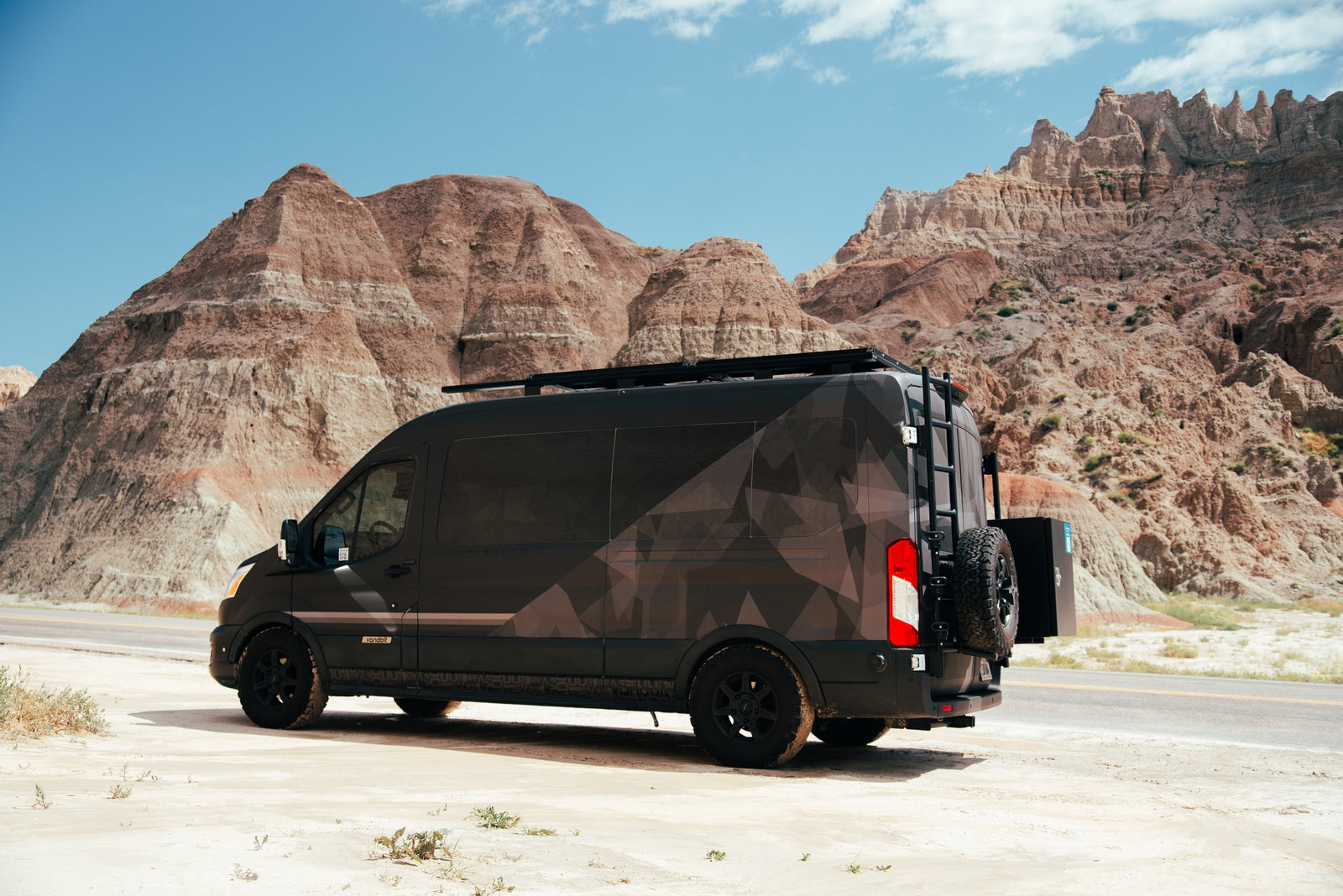 Customized dark RV rental van with a geo pattern and roof rack parked in rugged badlands terrain.