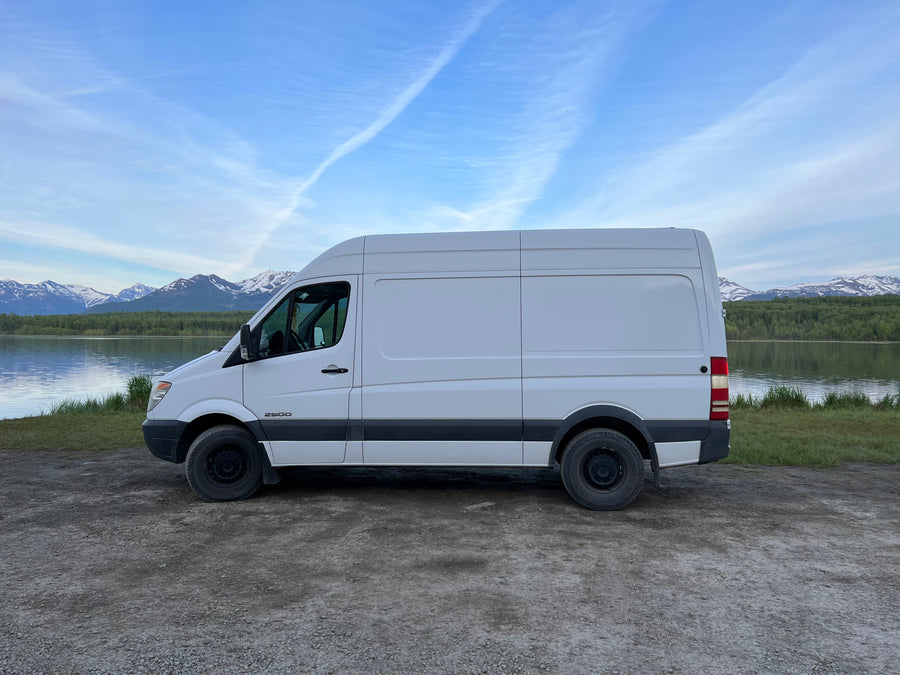 This white Sprinter van, ready for RV rental, is parked by a tranquil lake with snow-capped mountains.