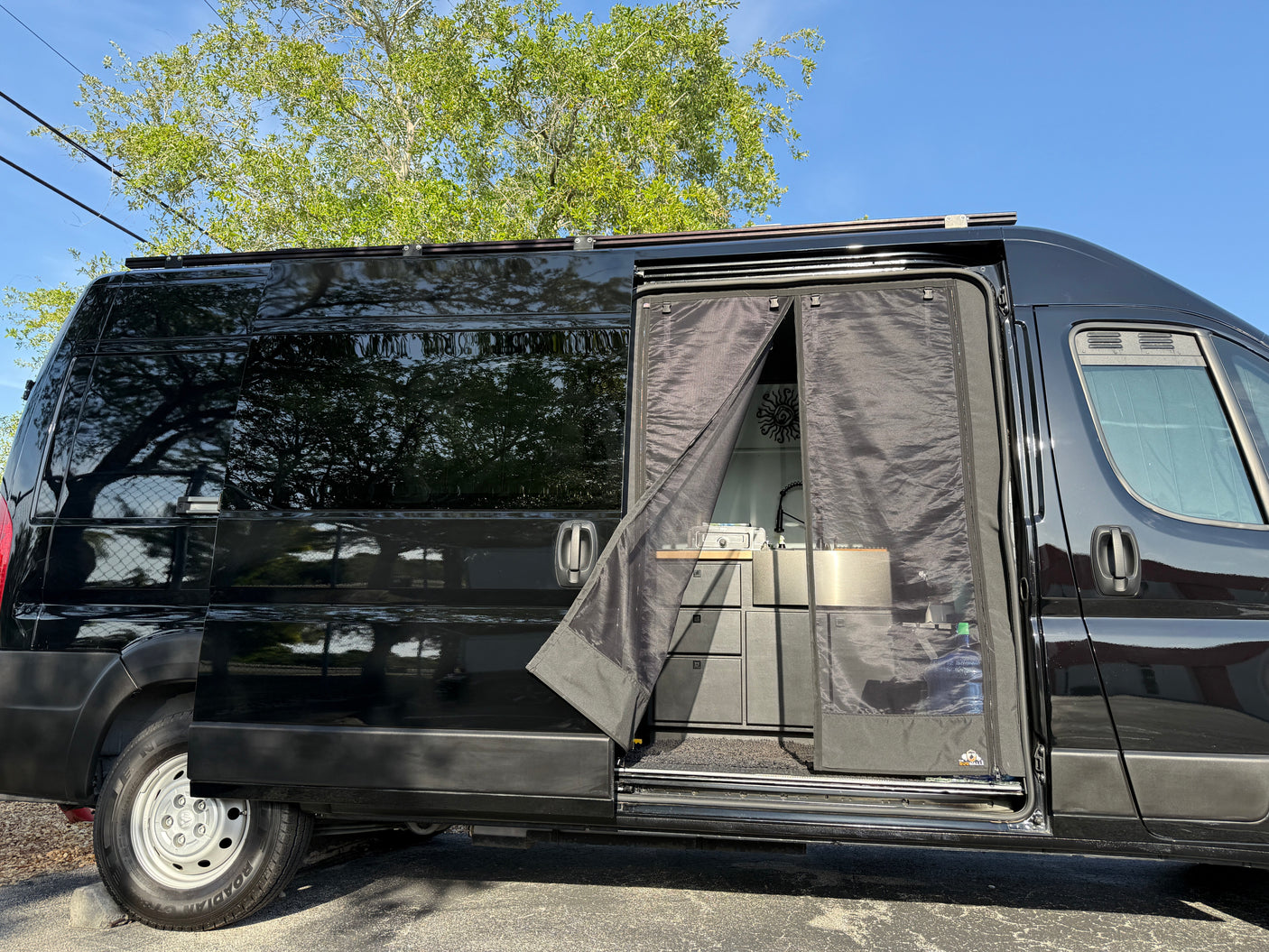 Black camper van for rv rental with open side door. Interior kitchen setup: sink, cabinets, & mosquito net visible.