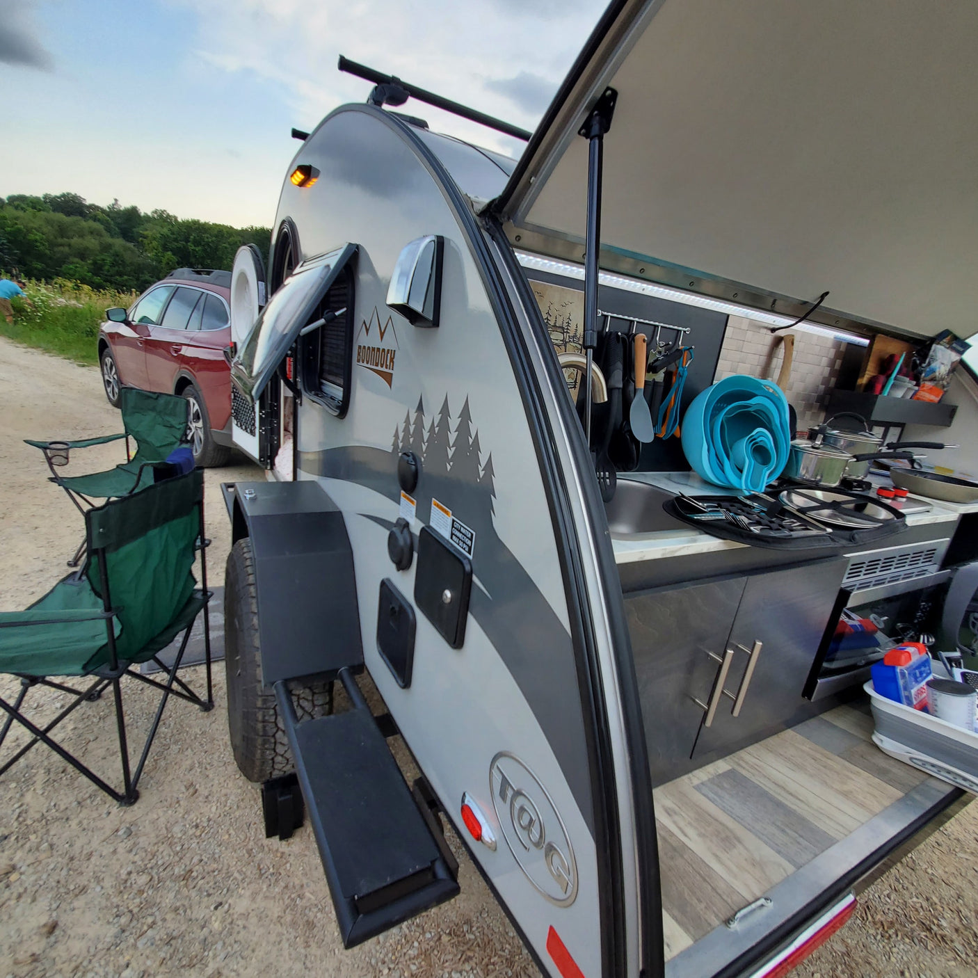 An open T@G Boondock teardrop camper's outdoor kitchen is set up with chairs for an rv rental camping trip.