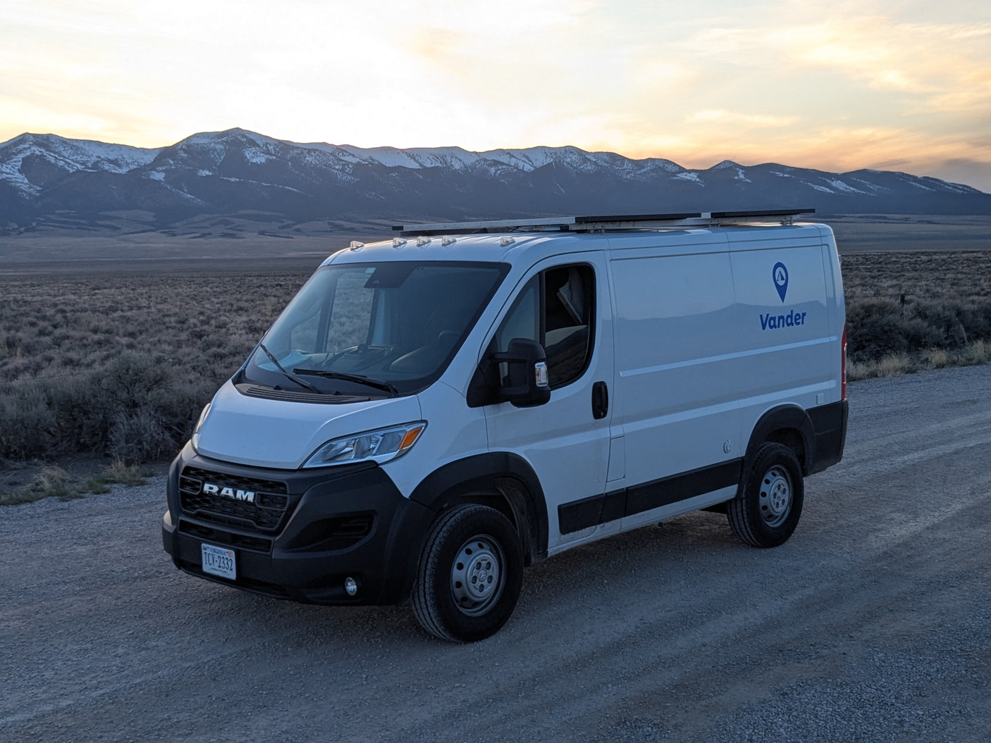 A white Vander RV rental van on a dirt road, backed by snow-capped mountains under a sunset sky.