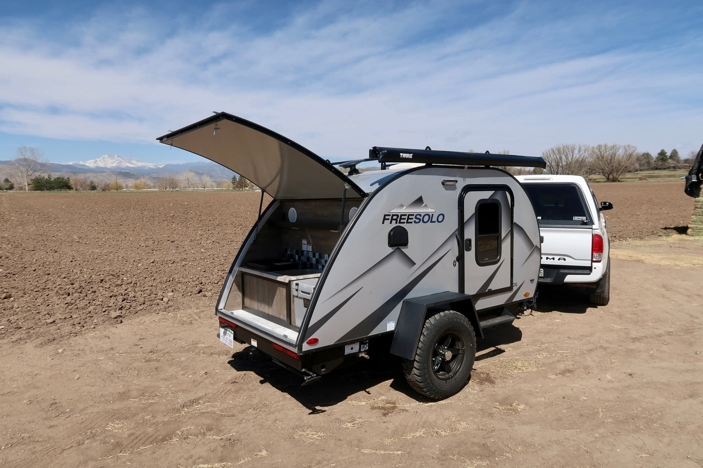 Gray FREESOLO teardrop camper (RV rental) with open galley & truck on dirt road. Mountains visible in background.