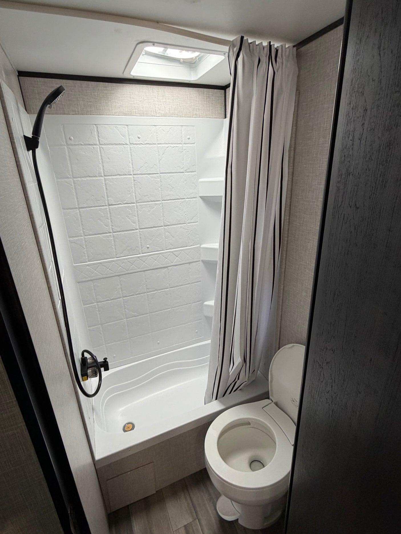 RV rental bathroom: white shower/tub, black striped curtain, overhead skylight, and toilet on gray floor.