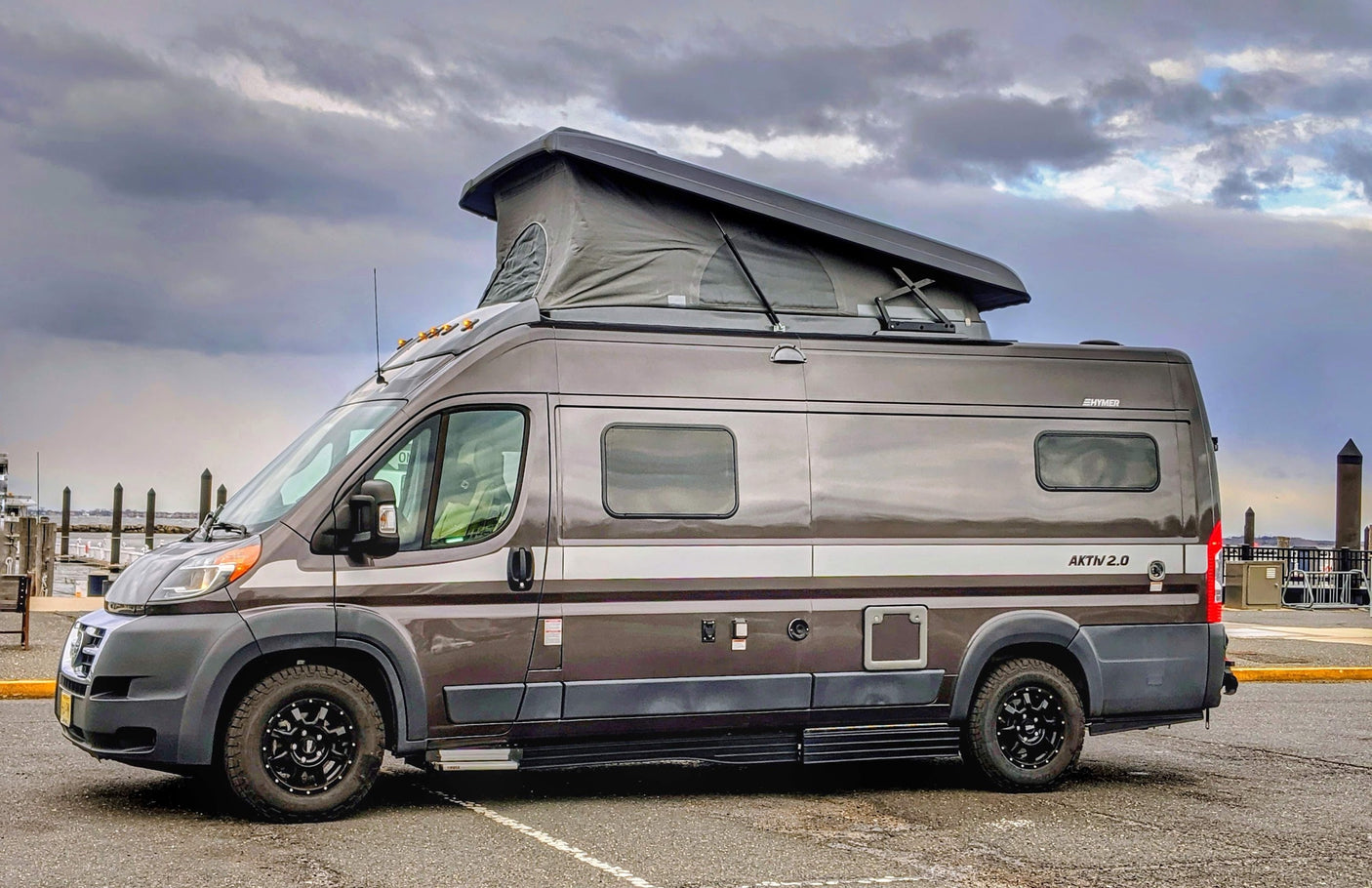 This sleek Hymer Aktiv 2.0 pop-top camper van, ideal for rv rental adventures, is parked by a cloudy waterfront.