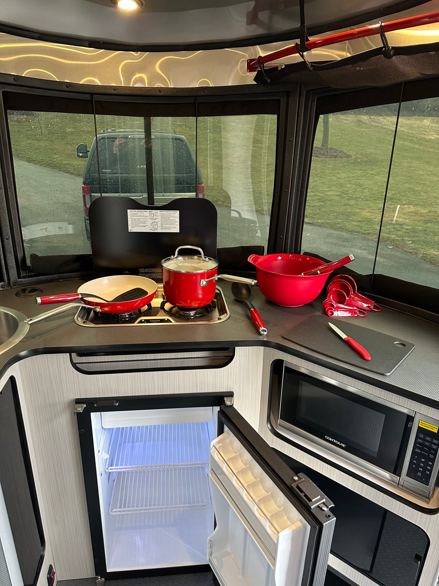 Cozy RV rental kitchen features stove, microwave, open fridge, and red cooking utensils, with a rainy outdoor view.