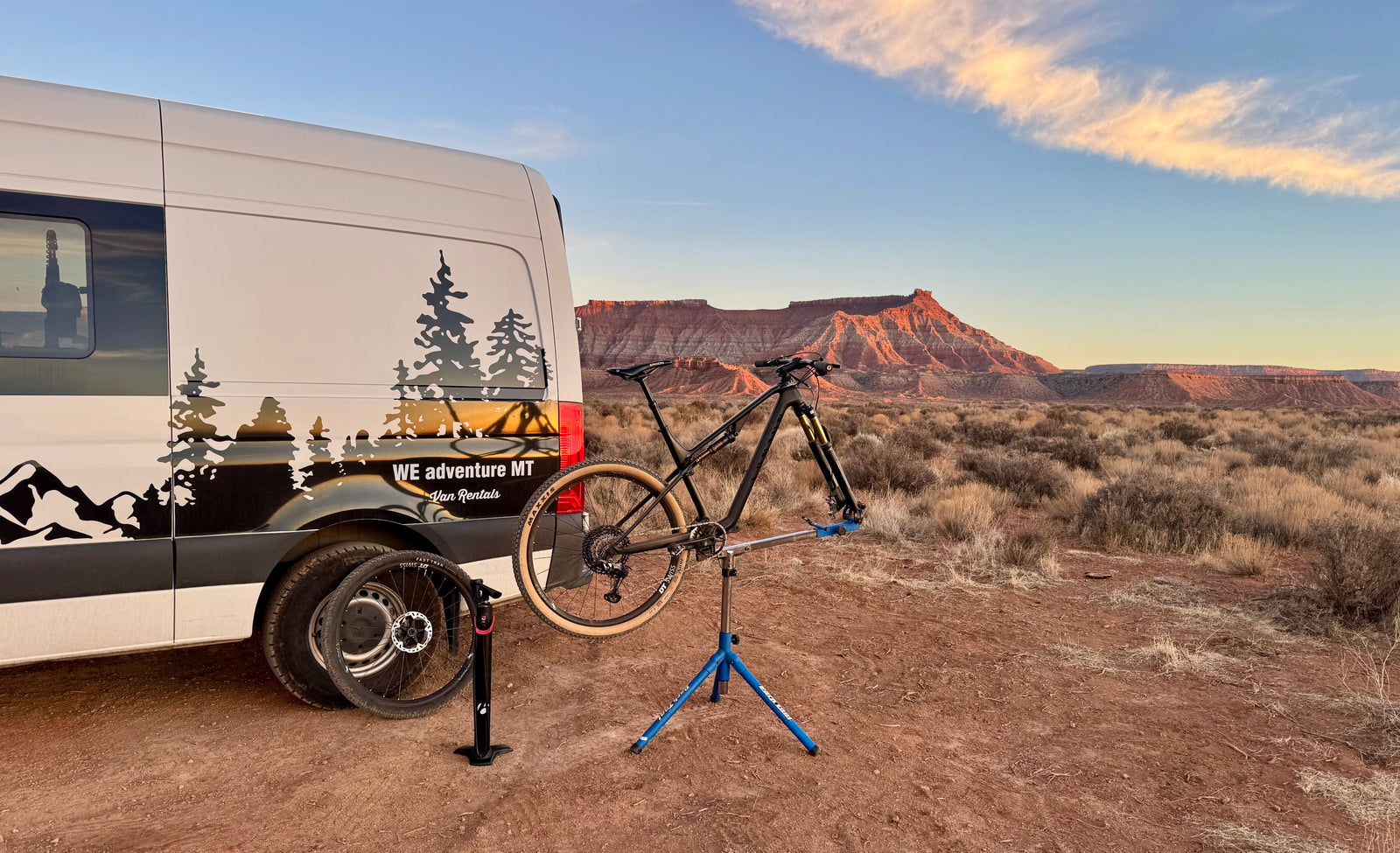An RV rental van and mountain bike being serviced on a stand, set against a desert sunset.