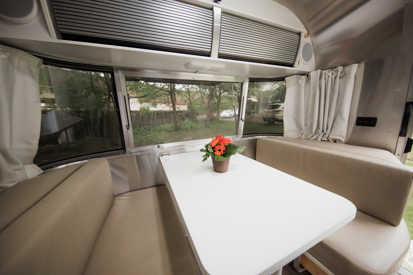 Inside this RV rental: a bright dinette with panoramic windows, comfy beige cushions, and a white table with flowers. Enjoy the scenic views!
