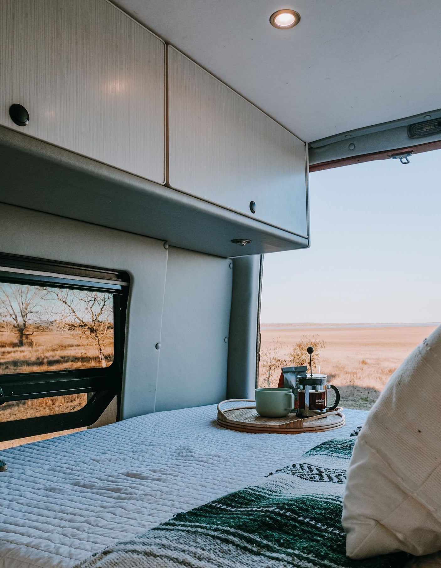 Inside an RV rental: a cozy bed with coffee on a tray, framed by windows showing a vast, dry landscape. Ideal for travel.