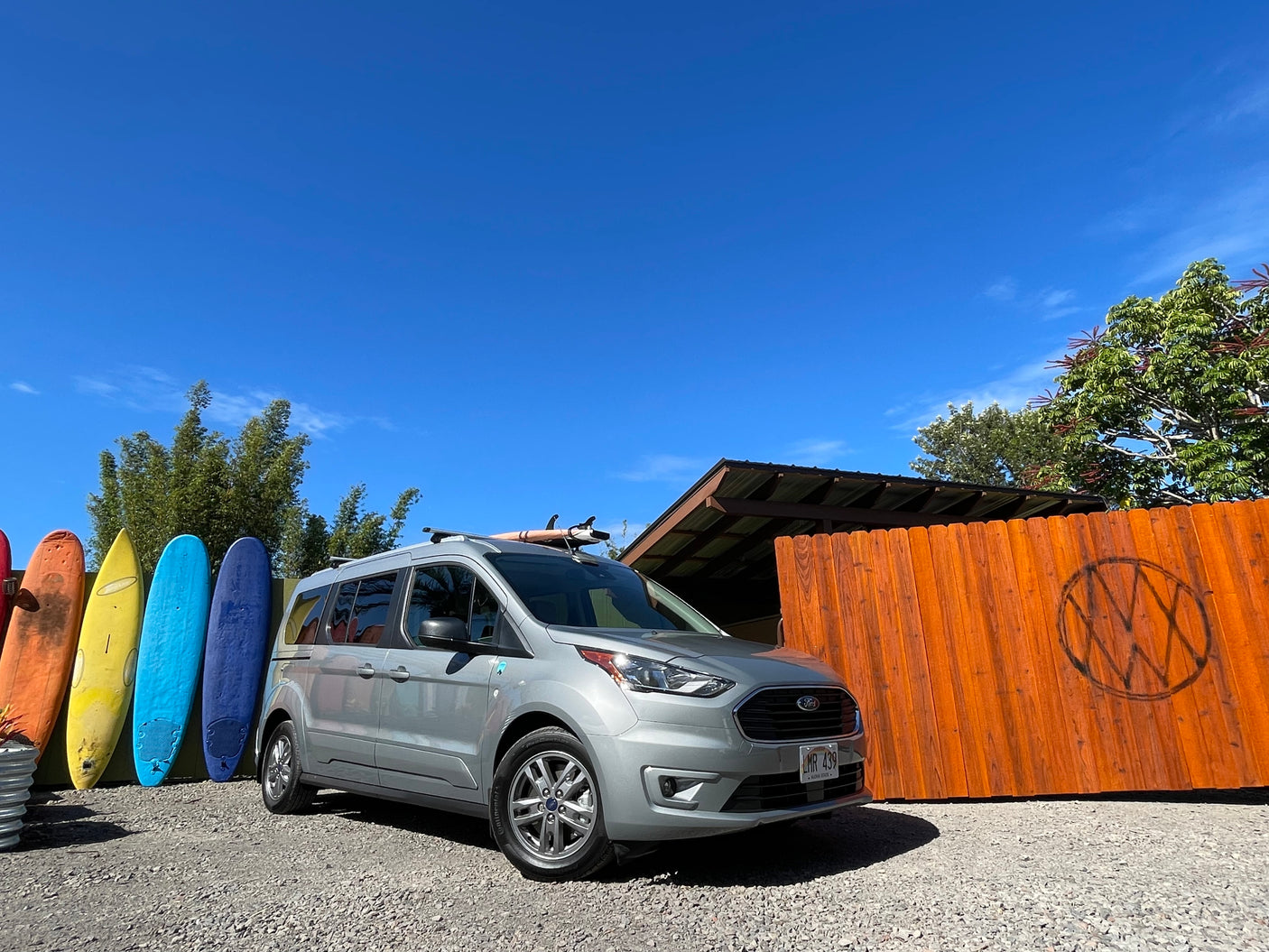 Silver Ford Transit Connect rv rental with surfboard on roof, parked by colorful surfboards & wooden fence under a blue sky in Hawaii.