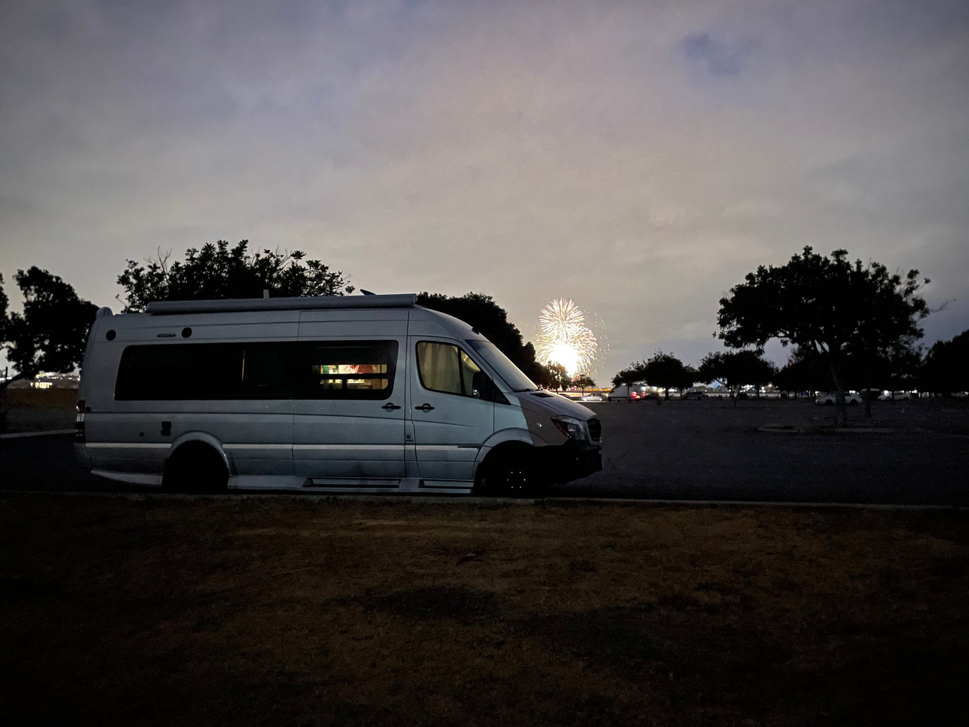 Dusky sky over a gray camper van RV rental with bright fireworks in the distance.
