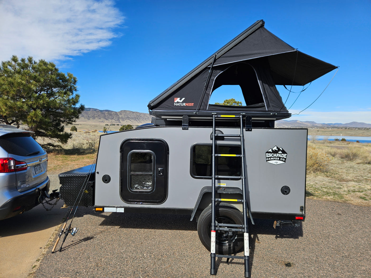 RV rental setup: Grey Escapade camper trailer with NaturNest rooftop tent & ladder, hitched to a Subaru. Ready for outdoor adventure.
