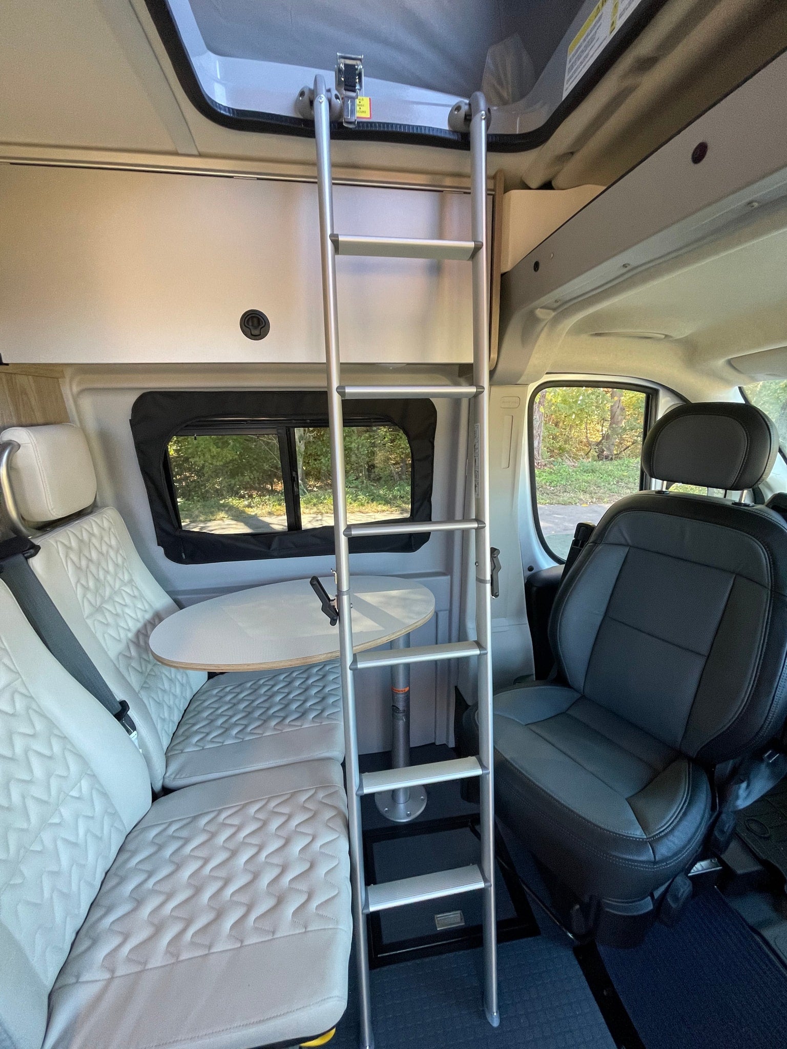 RV rental interior view: ladder to an overhead bed, dinette with light upholstery, and a dark gray driver's seat.