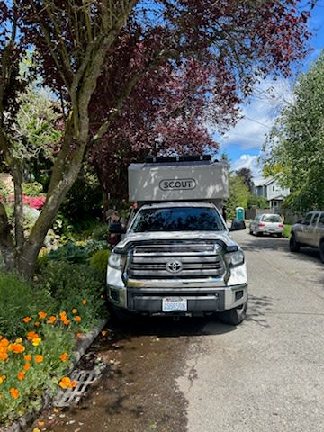 White Toyota pickup with a Scout truck camper, an RV rental, parked on a residential street with flowers.
