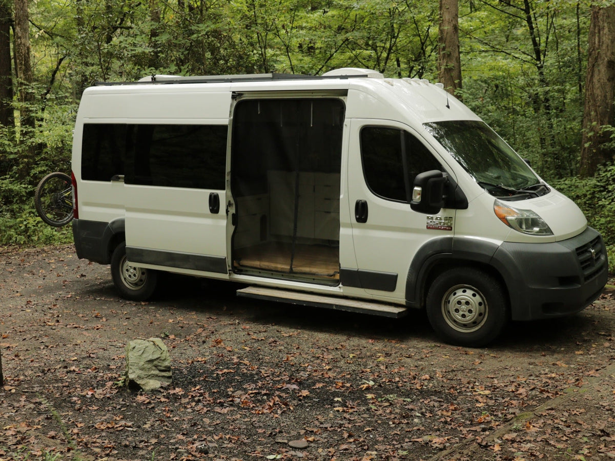 White Dodge ProMaster camper van (RV rental) parked in a leafy forest. Side door open — photo 1