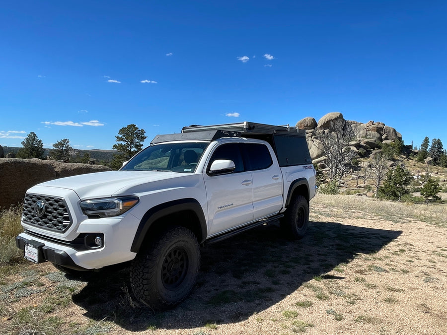 White Toyota Tacoma truck with an overland camper setup, parked off-road in a scenic, rocky area. An adventurous alternative to standard rv rental.