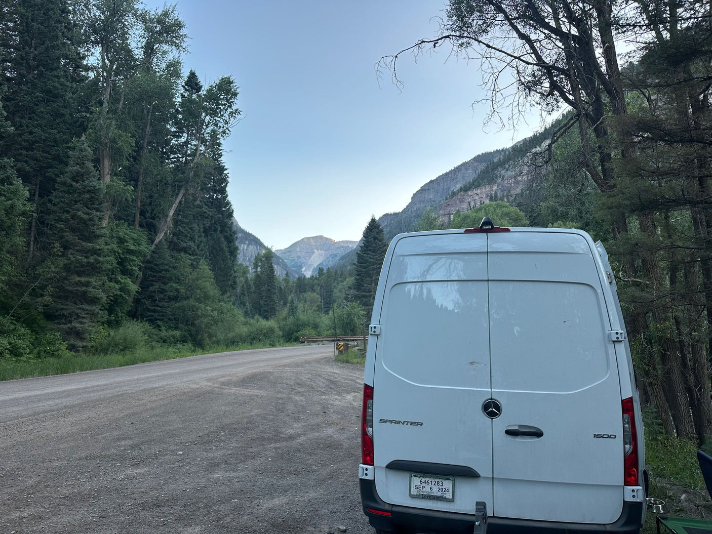 Adventure awaits! White RV rental van parked on a dirt road in a scenic mountain valley, surrounded by forests.