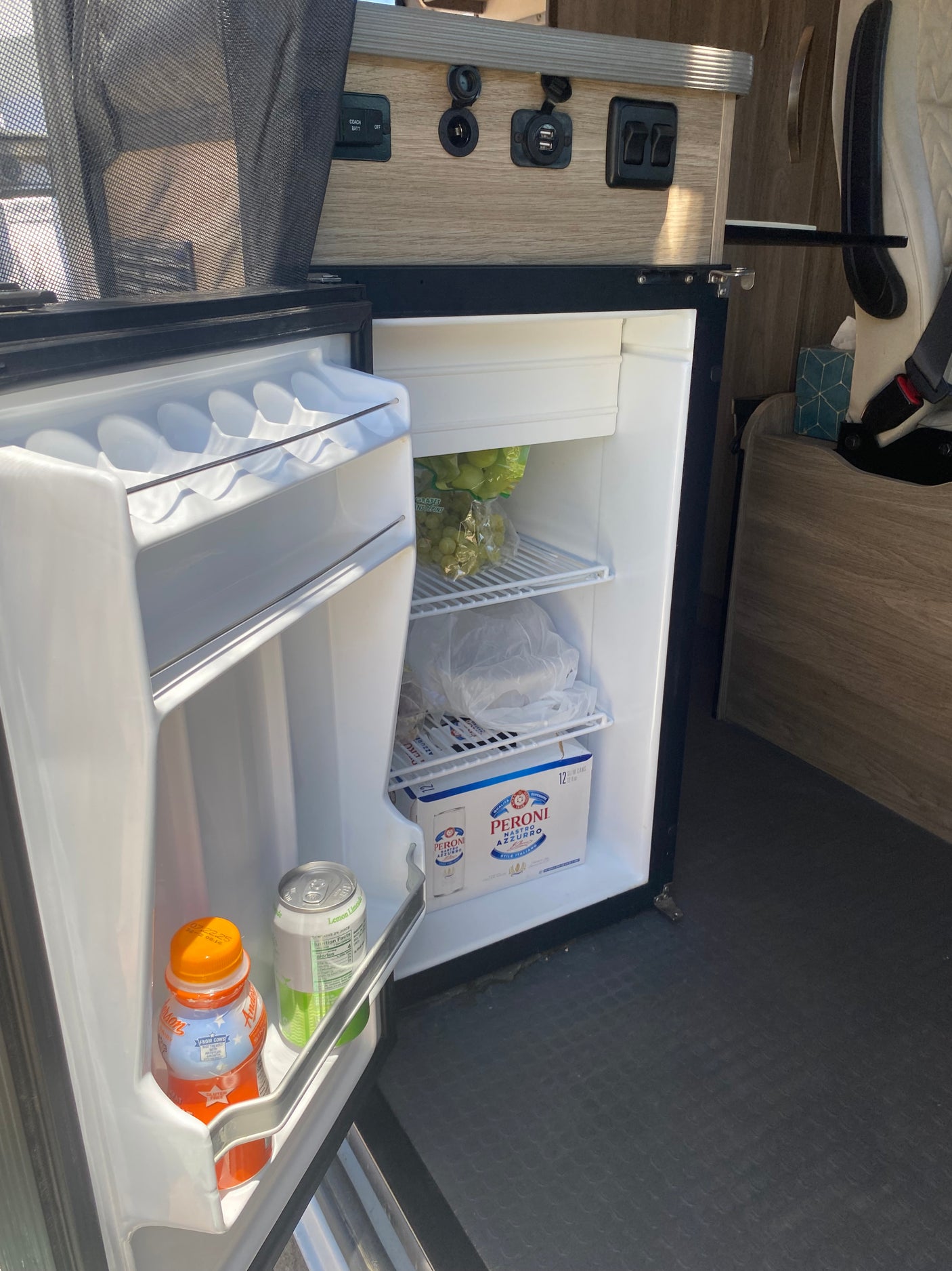 A compact, stocked mini-fridge inside an RV rental, featuring grapes, assorted drinks, and Peroni beer.