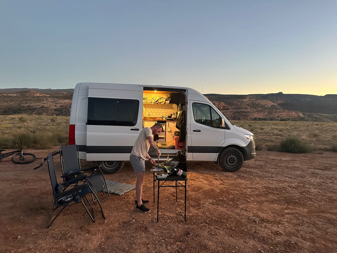 A man cooks dinner next to a white RV rental camper van at sunset in a desert landscape.