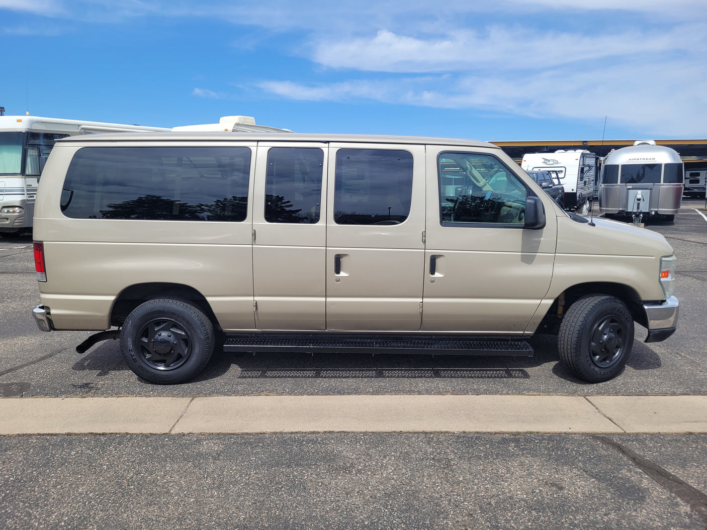 This tan Ford van is parked among various RVs, including an Airstream trailer, possibly for an RV rental business.