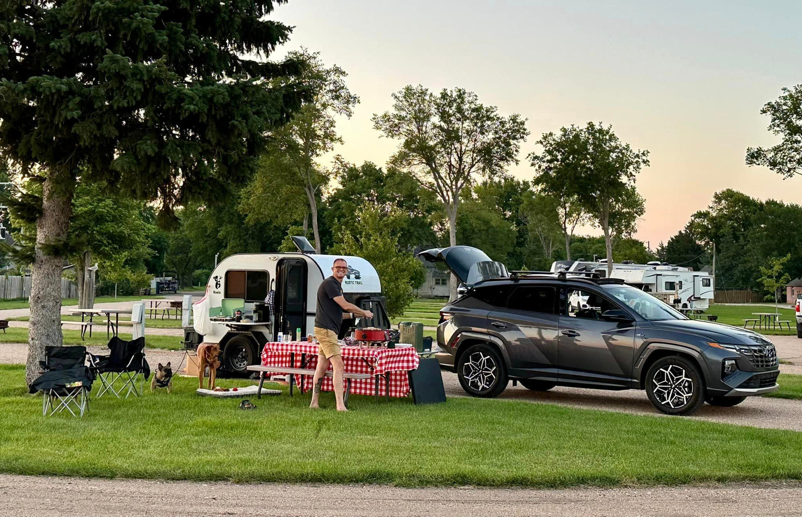 Enjoying an RV rental experience: man grilling outside a teardrop camper with dogs and SUV at a peaceful campsite.
