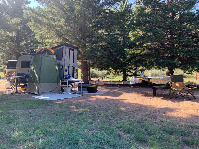 Relaxing RV rental campsite. Features a pop-up trailer with a privacy tent, picnic table, and chairs shaded by tall trees.