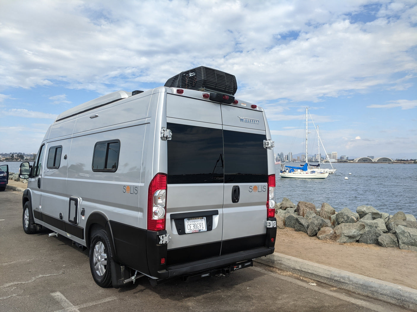 Experience travel with this RV rental! A silver Winnebago Solis camper van parked by the water, with boats and city views.