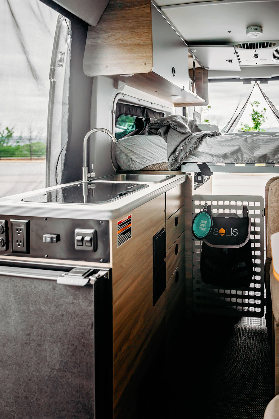 RV rental: Inside a modern camper van. Features a compact kitchen with sink, fridge, controls, and an elevated bed.