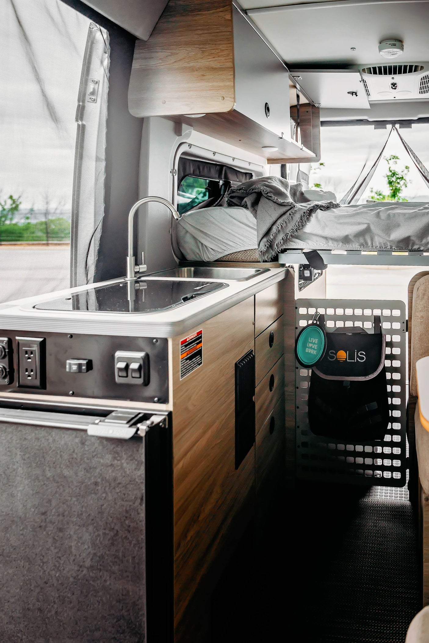 RV rental: Inside a modern camper van. Features a compact kitchen with sink, fridge, controls, and an elevated bed.