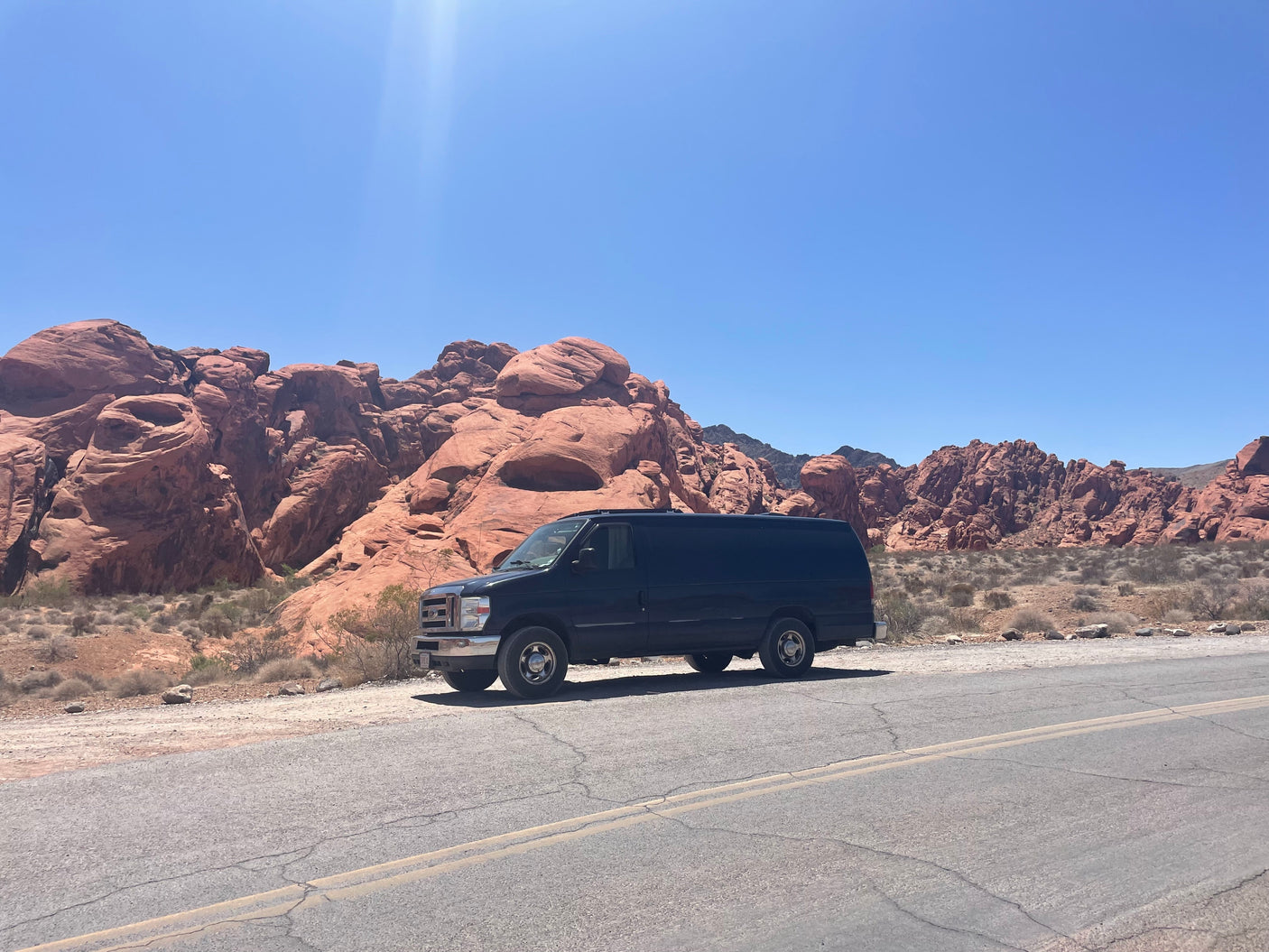 A black van on a desert road by red rock mountains under a bright blue sky. Perfect for an RV rental adventure!
