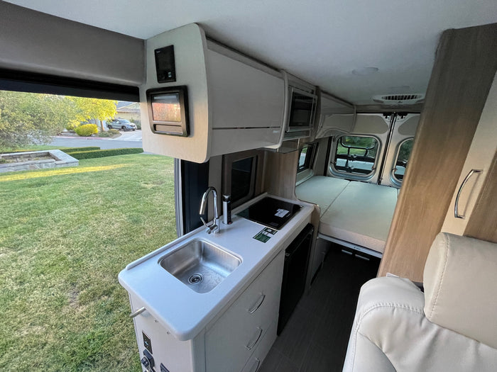 Inside an RV rental, showing the compact kitchen with sink, induction cooktop, rear bed, and outdoor view.