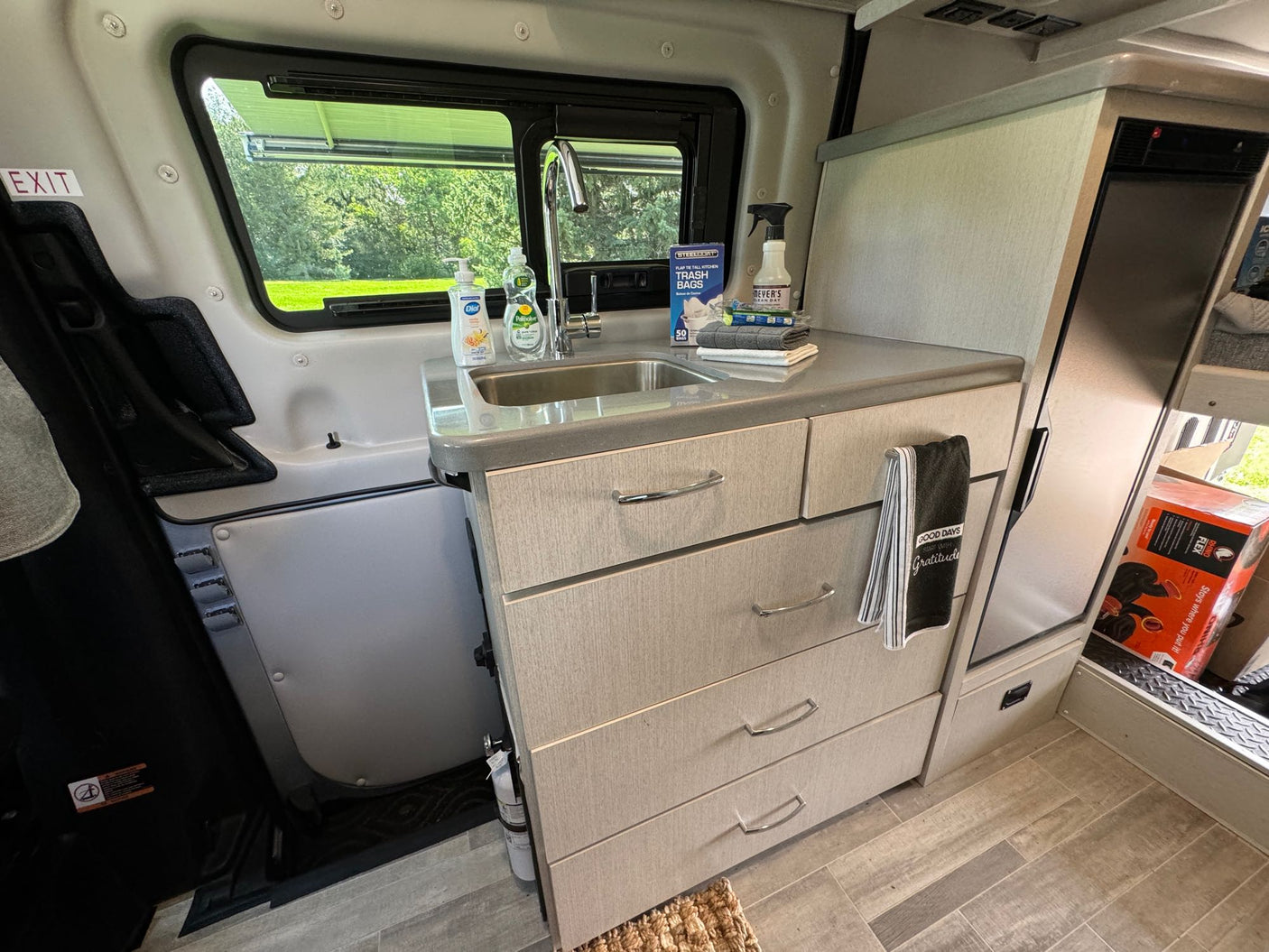 Inside an RV rental, a compact kitchen features a sink, countertop, multiple storage drawers, and a window view of green trees.