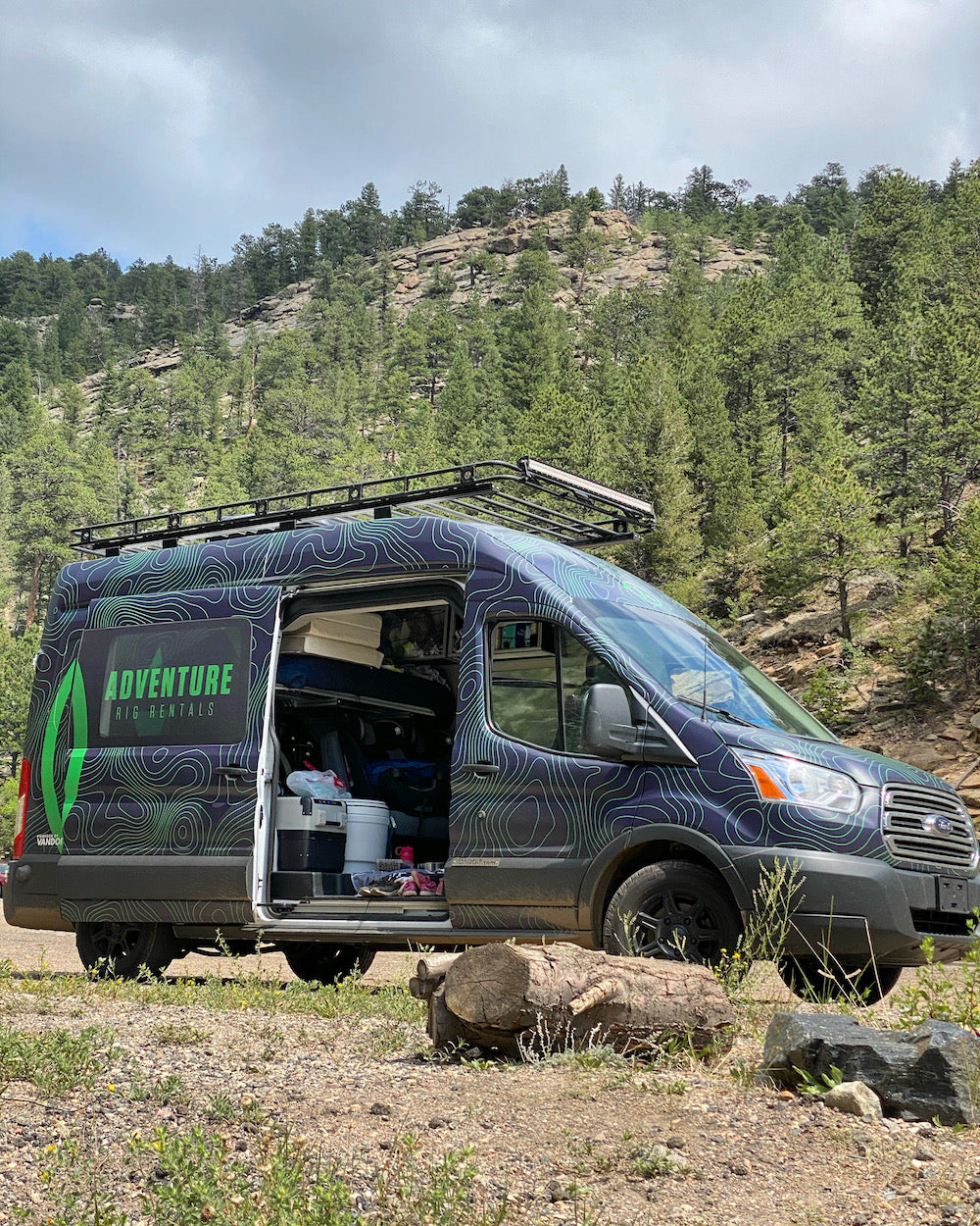 Dark-colored adventure RV rental van with open side door, parked in a forest, showcasing its camper features.