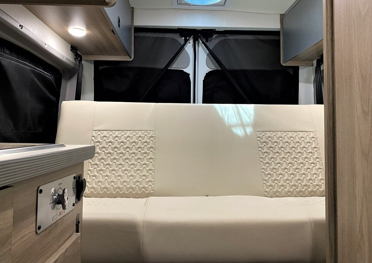 Clean RV rental interior: A cream bench seat with textured details, overhead lights & cabinets, & privacy-covered rear windows.