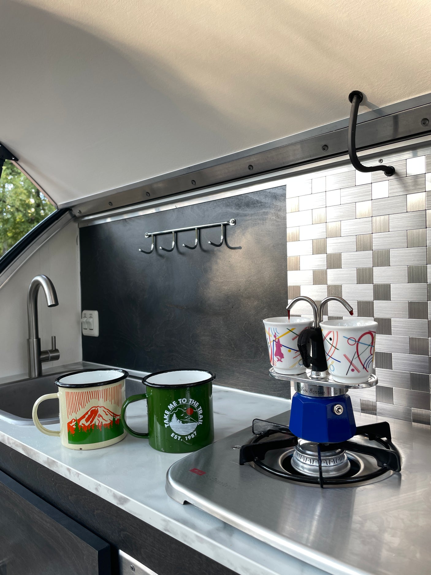 Fresh coffee brews in a Moka pot on a stove in a stylish RV rental kitchen, with two enamel mugs waiting.