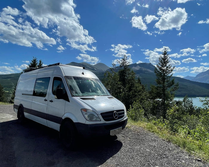featuring a side ladder & roof setup.|A white rv rental van is parked on a scenic mountain road overlooking a tranquil lake under a blue