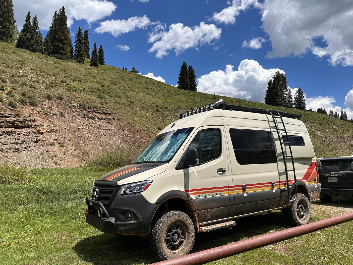 Storyteller Overland van, an adventure RV rental, parked on a grassy hill with tall trees under a bright sky.