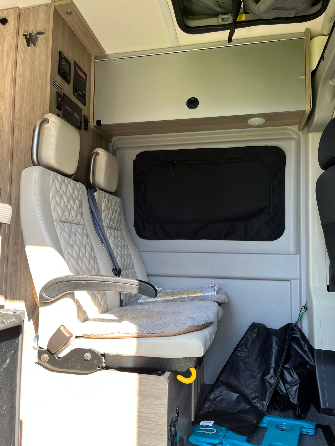 Inside an RV rental: light, quilted seats, a black window shade, controls, & storage cabinets, ready for your journey.
