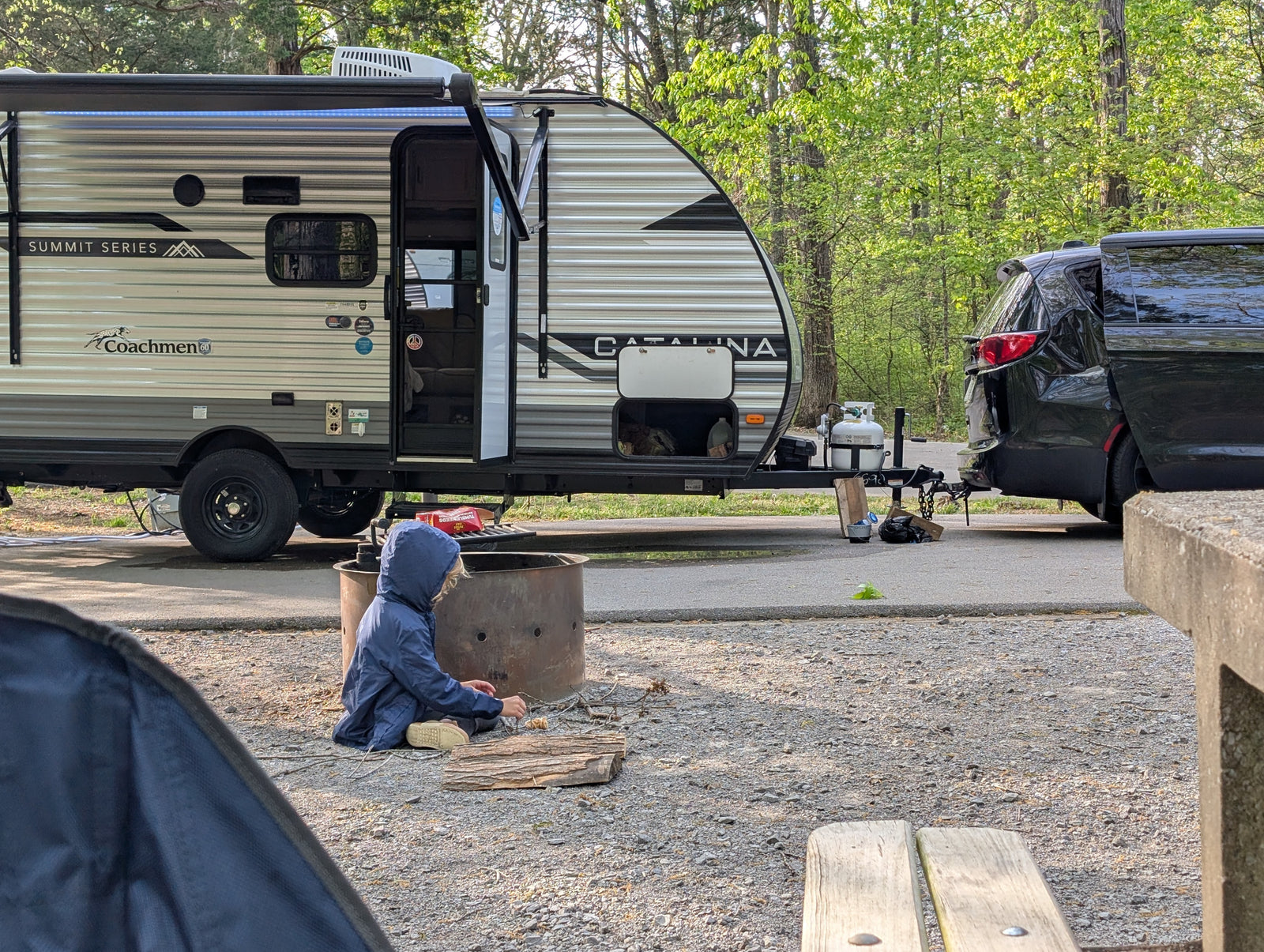 A child prepares kindling by a fire pit at a Coachmen Catalina travel trailer RV rental campsite.